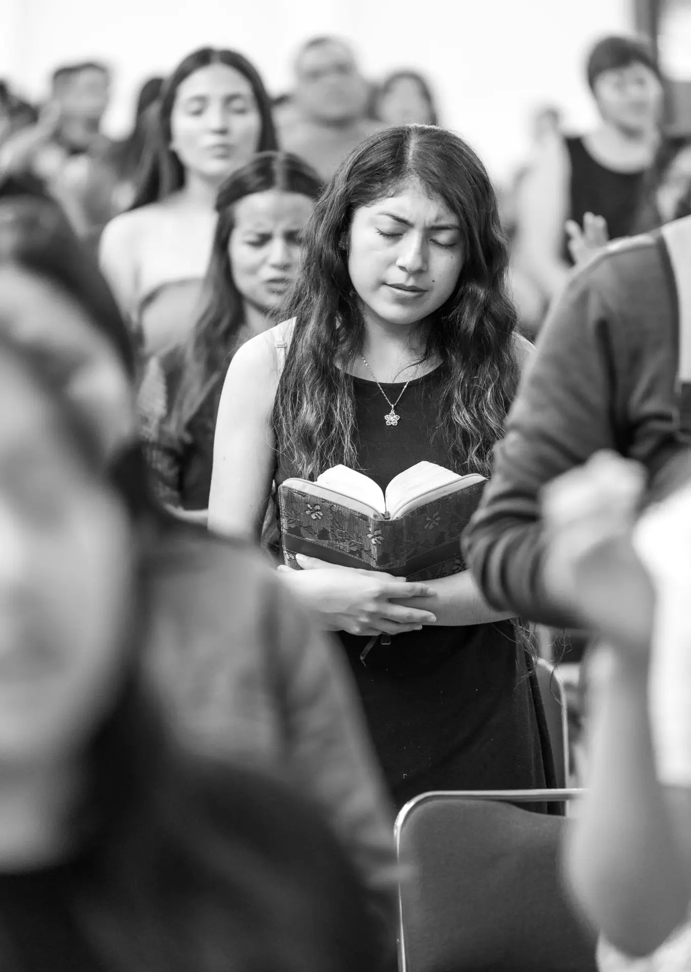A woman stands in prayer with a Bible in a church setting, eyes closed in devotion.
